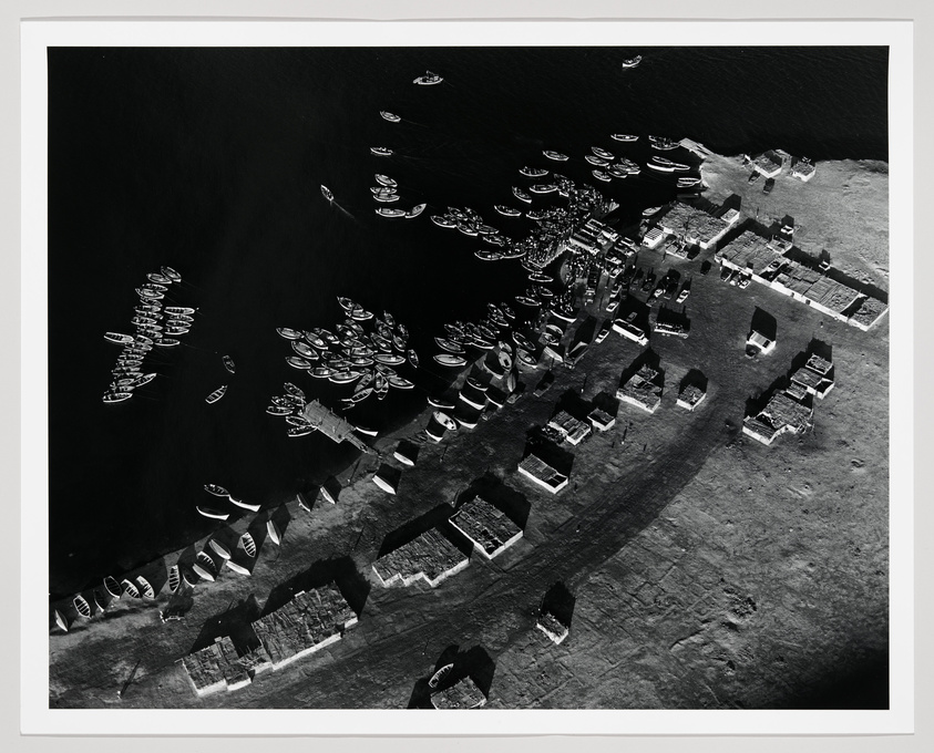 Aerial black and white photograph showing numerous boats clustered near a pier with several buildings adjacent to the waterfront. The contrast between the dark water and the lighter land creates a stark visual effect.