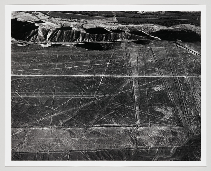 Aerial black and white photograph of the Nazca Lines in Peru, showing geometric lines and shapes etched into the desert landscape, with mountainous terrain in the background.