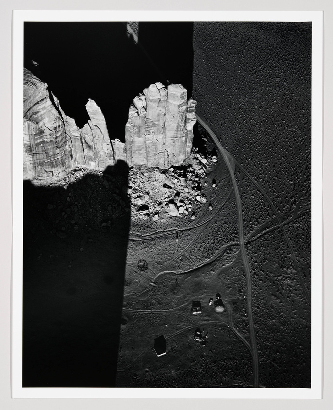 Black and white aerial photograph showing a rugged landscape with textured cliffs on the left and a smooth, dark surface on the right. A network of tire tracks crisscrosses the terrain, and several small, box-like structures are scattered across the bottom of the frame. The contrast between the rocky formations and the dark ground suggests a stark, desolate environment.