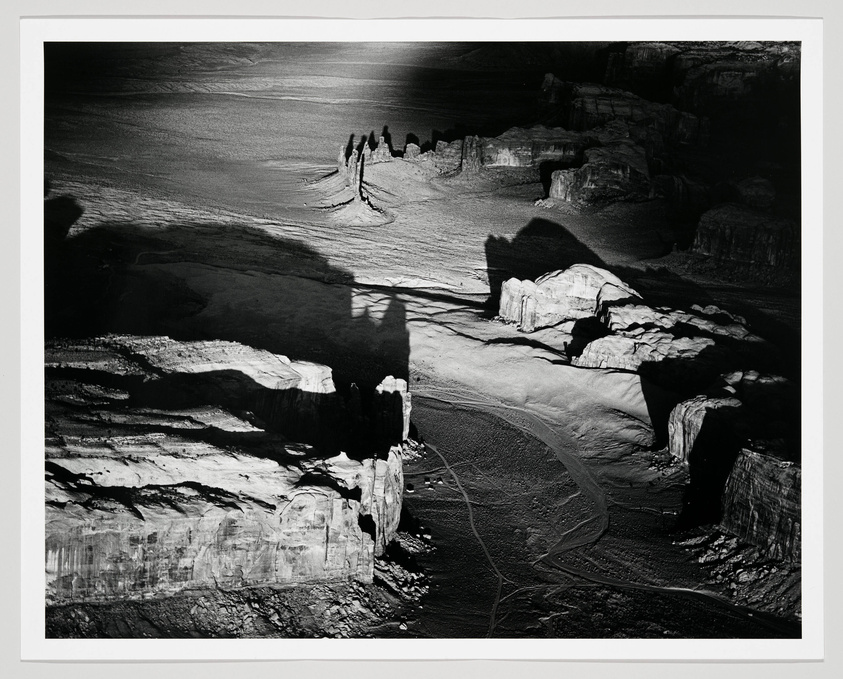 A black and white photograph capturing a dramatic landscape with deep shadows and contrasting highlights. The scene includes rugged terrain with large rock formations and striations, possibly a canyon, with a prominent shadow of a natural spire or monument cast onto the ground. Tire tracks are visible on the ground, suggesting recent vehicle activity. The image has a stark, almost otherworldly appearance due to the play of light and shadow.