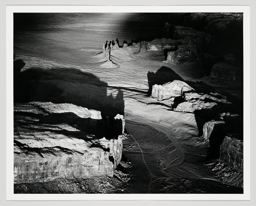 Aerial view of sunlit canyon cliffs casting long shadows over a winding sandy riverbed with vehicle tracks.