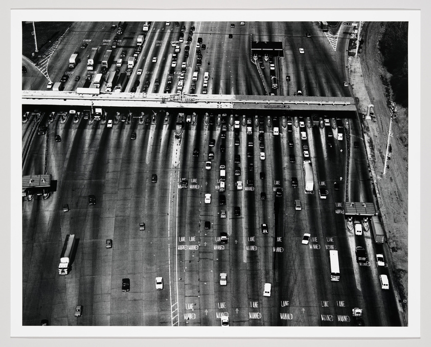 Aerial view of a multi-lane highway with heavy traffic, showing numerous cars and trucks in various lanes, some lanes marked as "LANE ENDS" and "MANNED," with a bridge overpass intersecting the highway. The image is in black and white, emphasizing the patterns of the vehicles and the roadway.