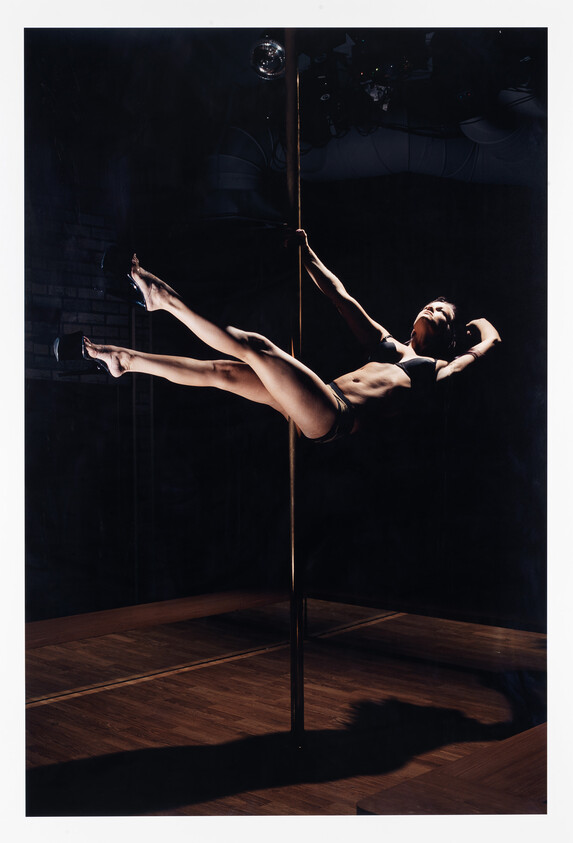 A woman holds a horizontal pole dance pose, gripping the pole with one hand and legs extended.