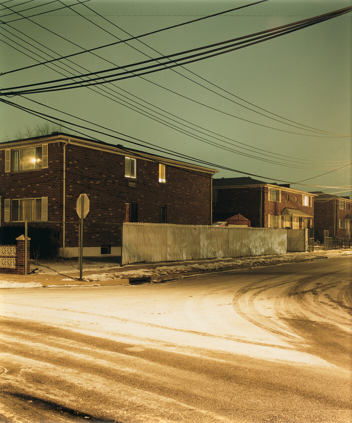 Brick apartment buildings along a snowy street under a web of overhead power lines at night.