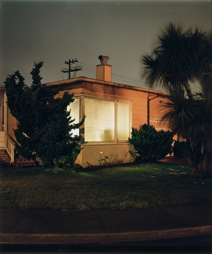 A nighttime view of a small, warmly lit house with a prominent front window. The house is painted in a light orange hue and has a chimney on the roof. In the foreground, there's a well-maintained lawn with several bushes and a palm tree swaying slightly in the breeze. The sky is dark, suggesting it's late in the evening.