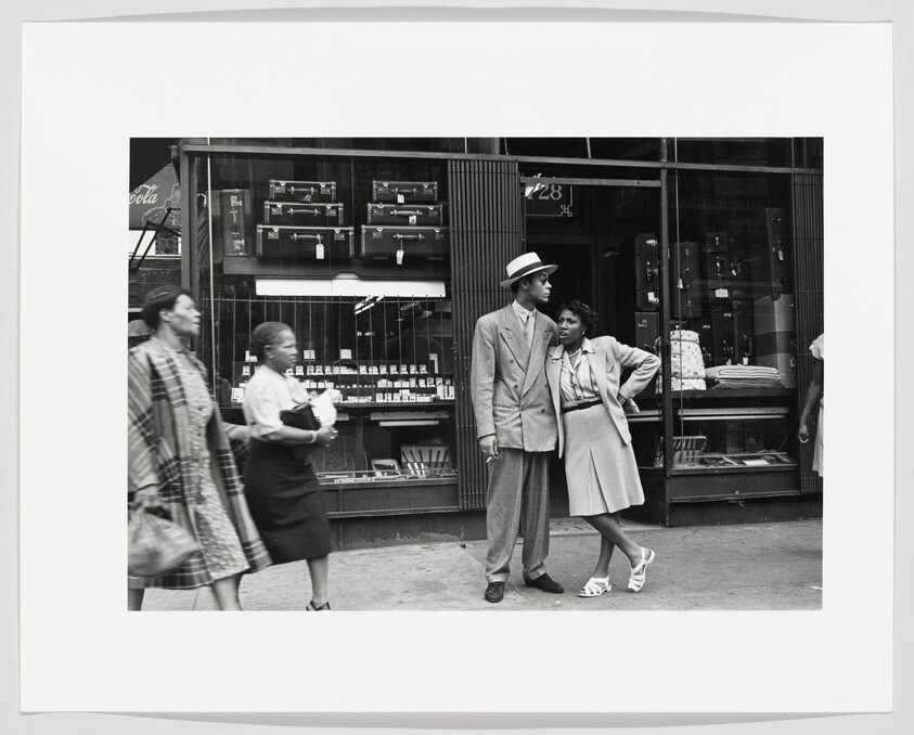 Young couple stands in front of a shop window, the woman leaning on the man's shoulder.