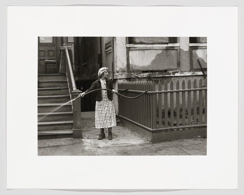 An older woman waters plants with a long hose beside a wooden picket fence.