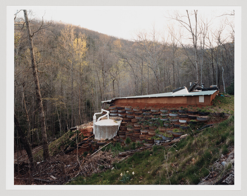 Small hillside house with stacked tire retaining wall and a white water tank connected by pipes.