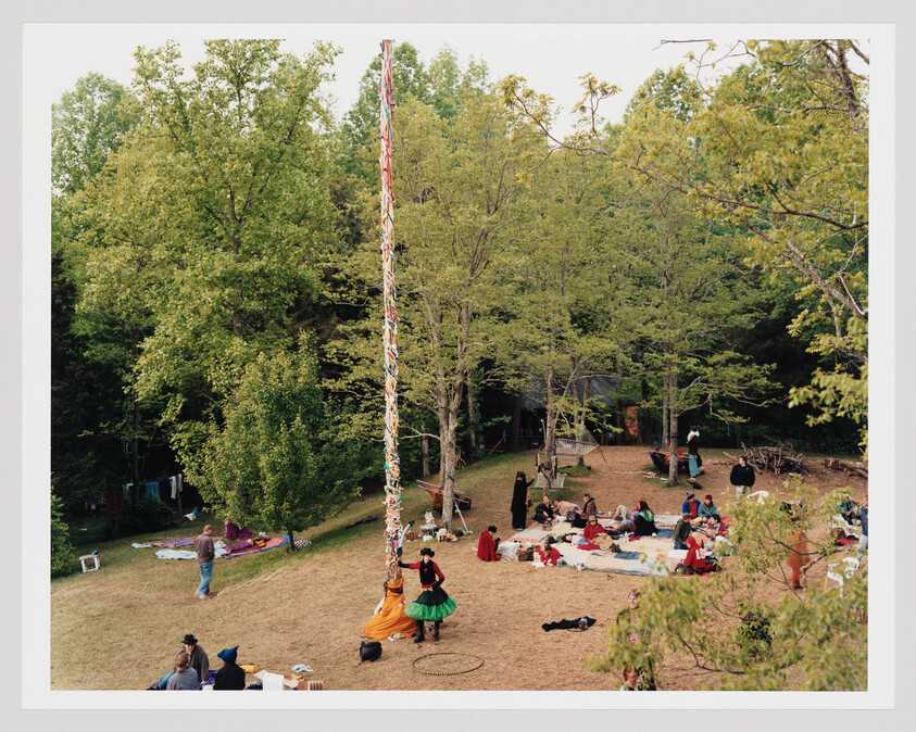 People gathered in a wooded clearing around a tall decorated maypole, with some dancing and sitting on blankets.