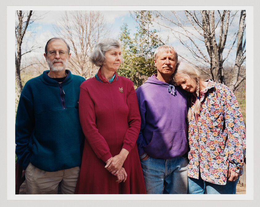 Four adults stand together outdoors, one woman resting her head on the shoulder of the man next to her.