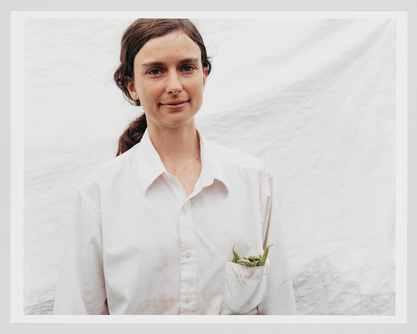 Young woman in a white shirt smiles with fresh peas tucked into her shirt pocket.