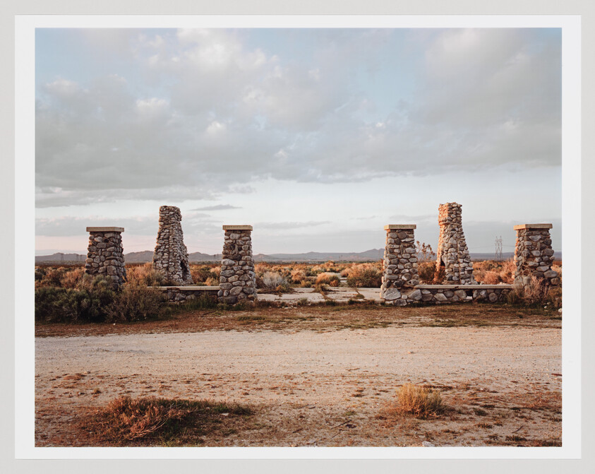 A row of six stone pillars forms an entryway in a dry desert landscape at sunset.