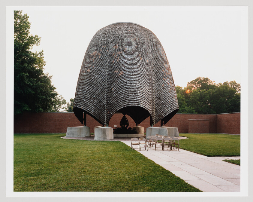 A large curved wooden-shingled pavilion stands over a small altar with folding chairs in a grassy courtyard.