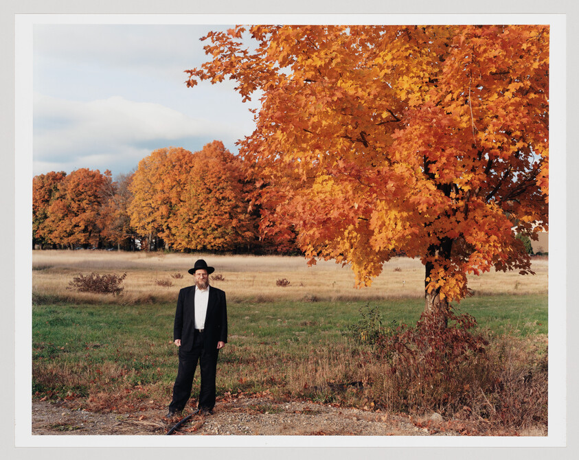 A man in a black suit and hat stands in a field beside a large orange autumn tree.