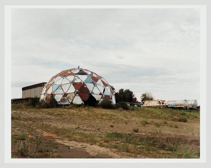A rusted geodesic dome with missing panels sits in an overgrown field beside industrial trailers.