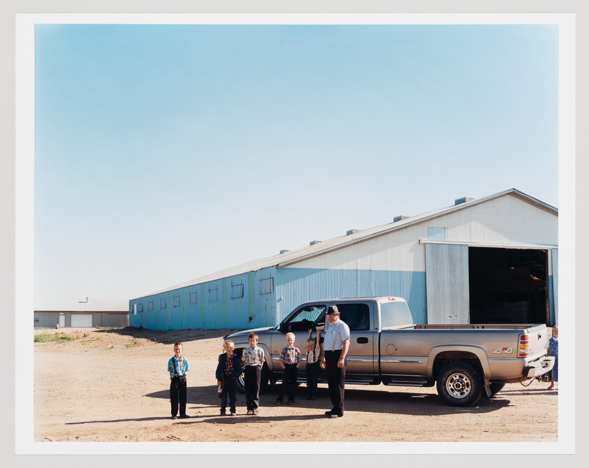 A man and five boys stand beside a silver pickup truck in front of a large blue warehouse.