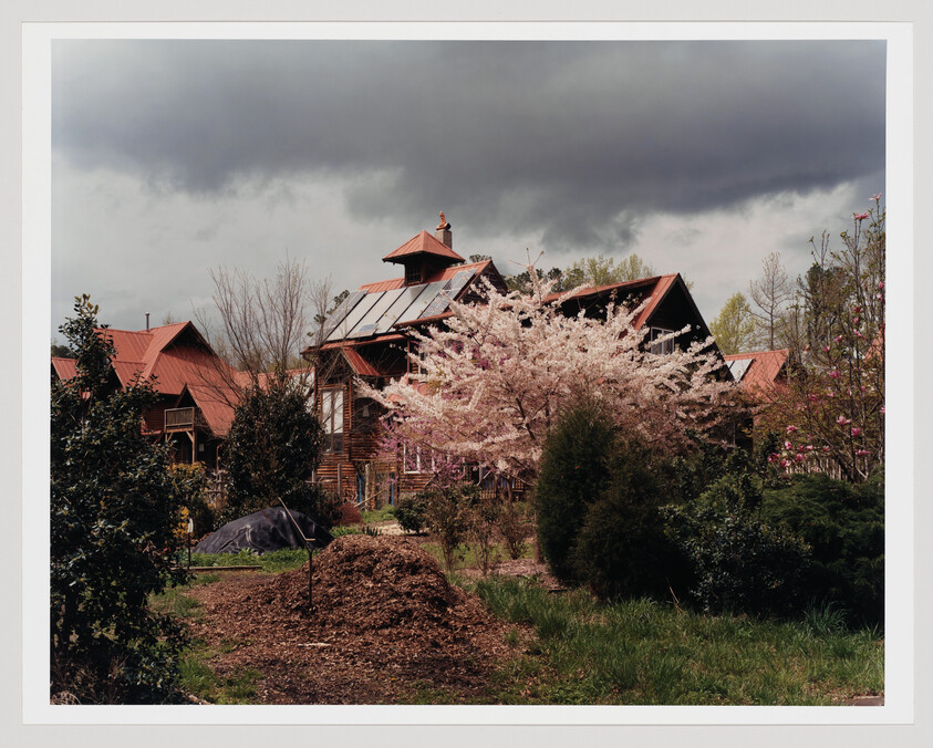 A rustic house behind a blooming cherry tree under dark storm clouds with a mulch pile in front.