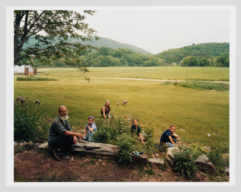 An older man and four children sit among garden flowers at the edge of a wide grassy field.