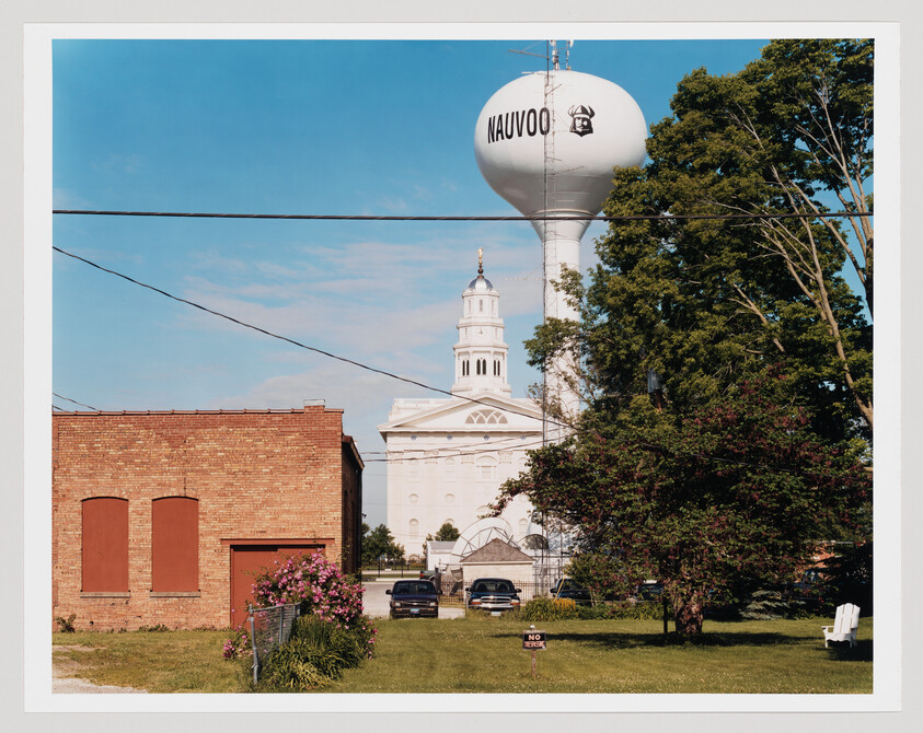 A tall water tower labeled "Nauvoo" rises behind a white church and brick building.