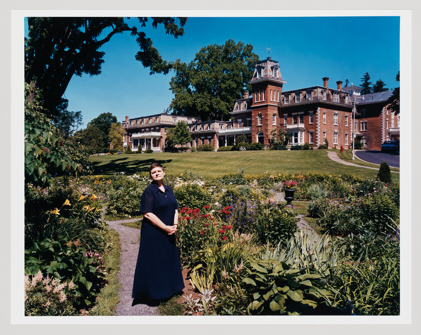 A woman stands on a garden path in front of a large historic brick mansion.