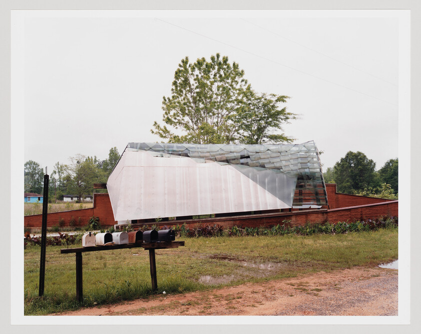 A row of mailboxes sits on a wooden stand in front of a curved metal and glass building.