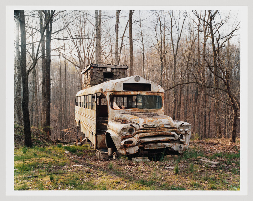 A rusted, abandoned school bus with a small wooden structure sits decaying in a leafless forest.