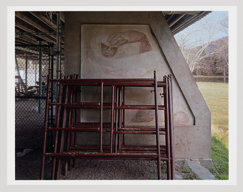 Stacked red scaffolding pipes lean against a concrete bridge pier with a faded mural.