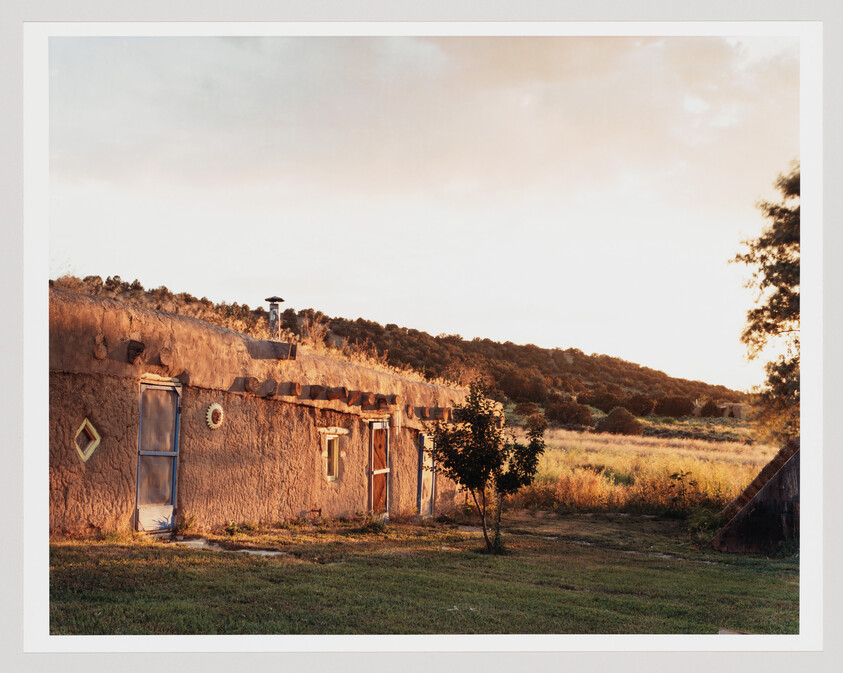 A traditional adobe house with a flat roof and wooden accents bathed in the warm glow of sunset, with a backdrop of a grassy field and trees under a soft sky.