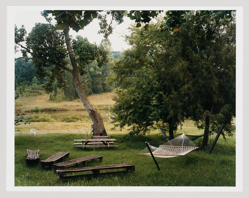 A hammock hangs between trees beside picnic tables and wooden benches on a grassy riverside.
