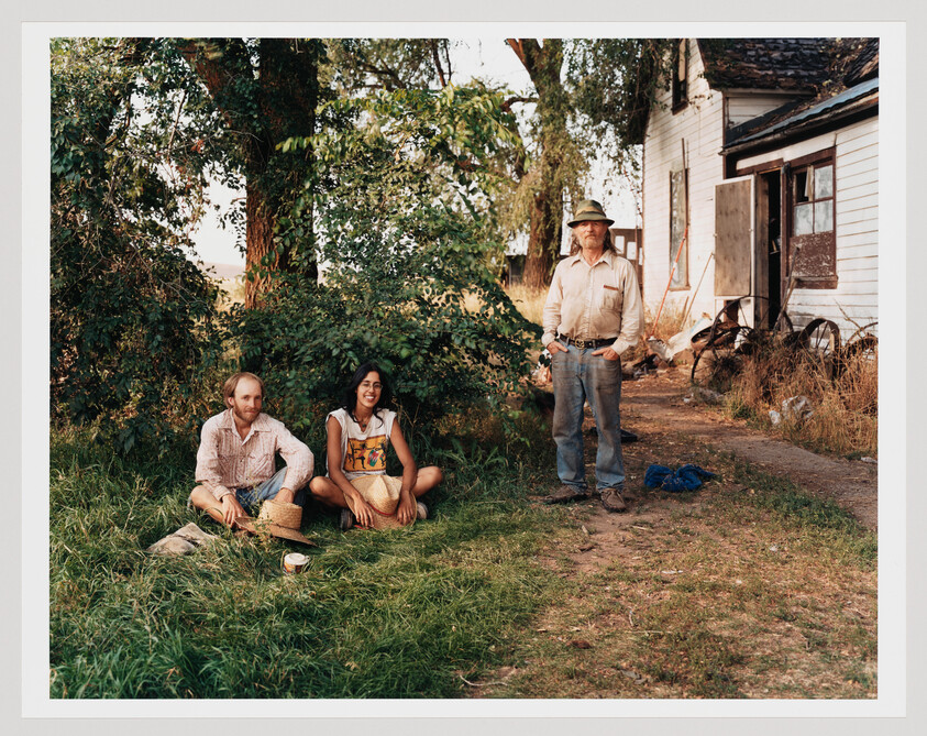 Two people sit on the grass under a tree while an older man stands by a weathered farmhouse.