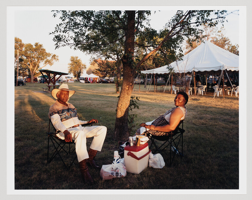 Two people sitting in folding chairs under a tree beside a cooler at a park event.