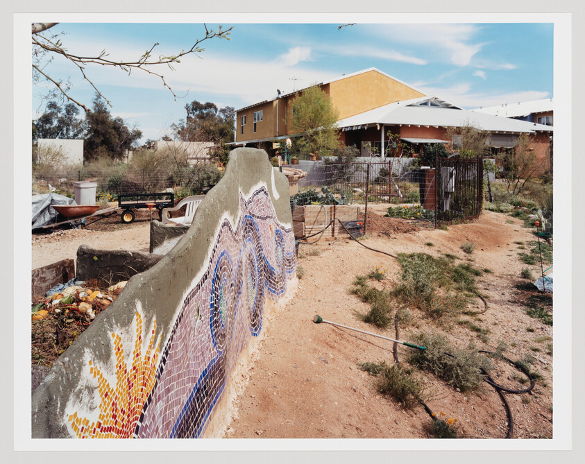 A mosaic-adorned low garden wall curves along a dry yard in front of a house.