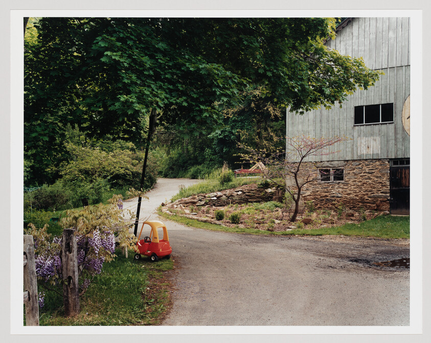 A red and yellow toy car sits beside a gravel driveway near a barn and trees.