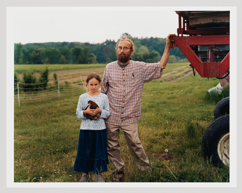 A bearded man stands beside a young girl holding a chicken in a grassy field.