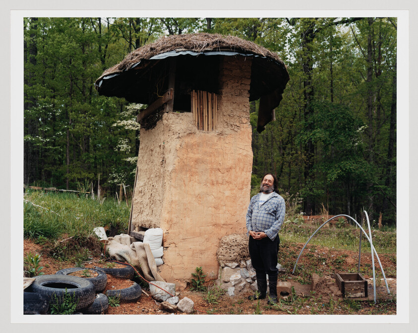 Man stands smiling beside tall cob house with thatched roof in wooded garden.