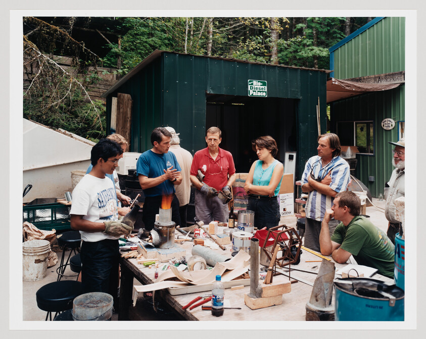 A group of people watch a metalworking demonstration around a cluttered outdoor workbench.