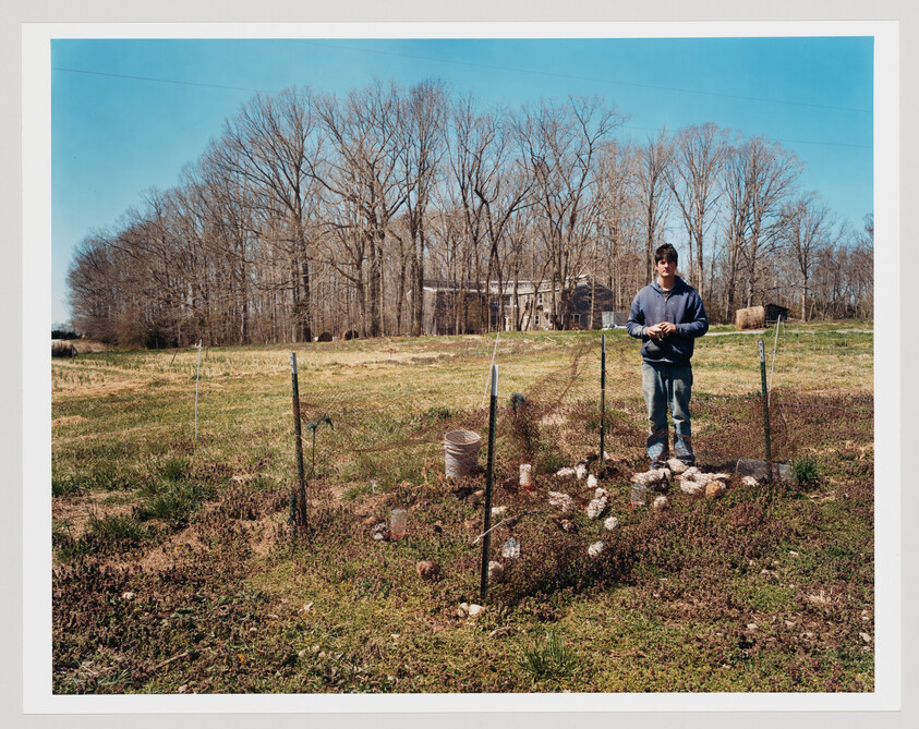 A young man stands inside a small fenced garden plot in a grassy field near trees.