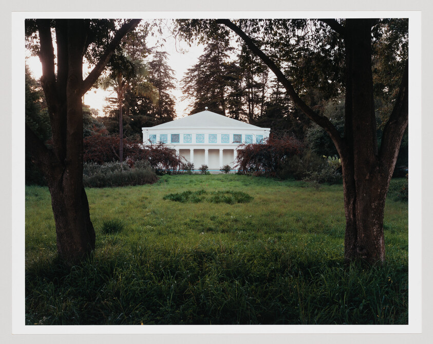 A white columned pavilion stands at the end of a grassy lawn framed by two trees.
