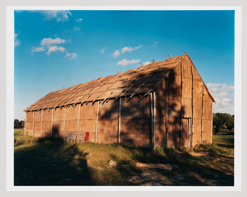 A large weathered wooden barn stands in a sunlit field with shadows and a row of chairs.