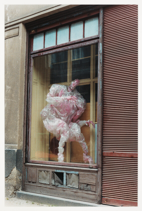 A translucent pink plastic humanoid sculpture is twisted and posed inside an old shop window.