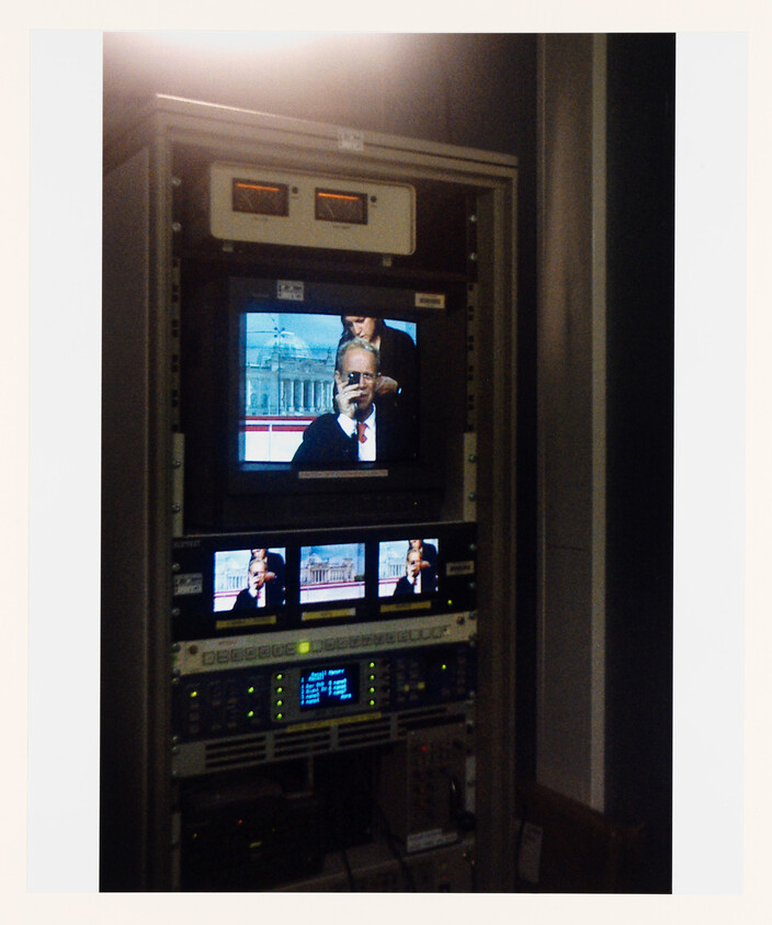 Television equipment rack with multiple monitors showing a man speaking on a news broadcast.
