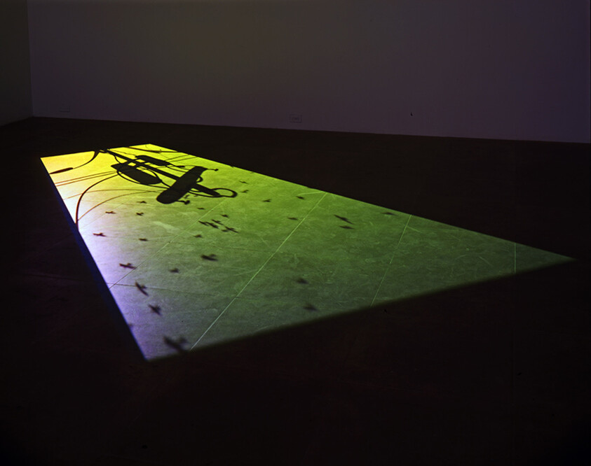 A projector casts a bright green light onto a floor, creating a rectangular shape with shadows of leaves and electrical cords visible within the illuminated area. The surrounding space is dimly lit, emphasizing the contrast between the projection and the dark floor.
