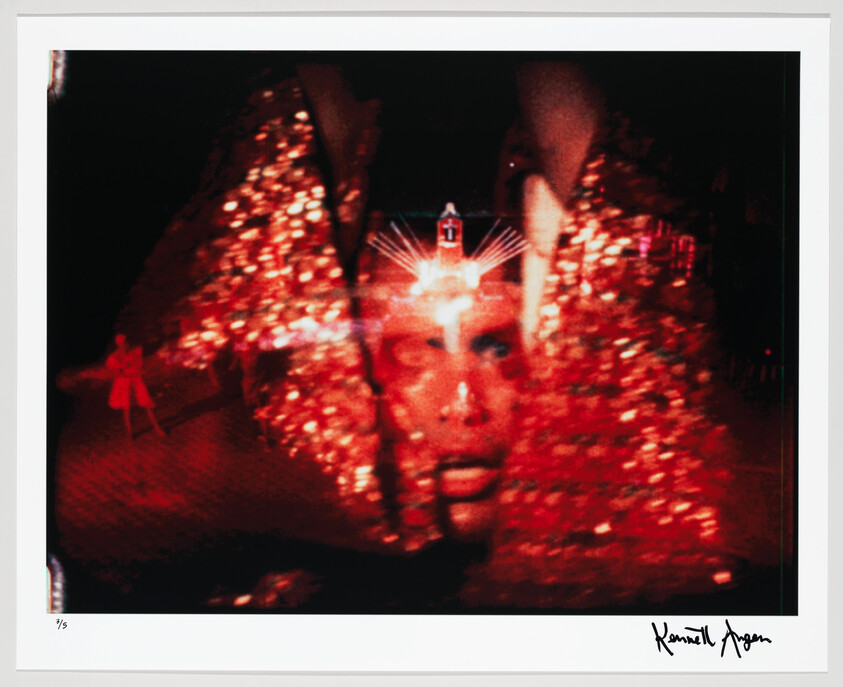 A glitter-covered performer’s face lit by red lights with hands framing the face.