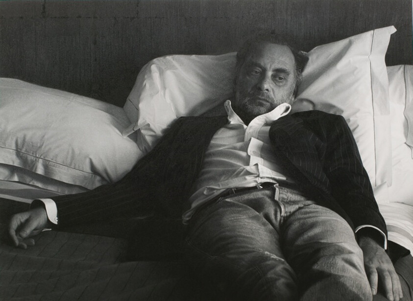 A tired middle-aged man in a jacket lies back on a bed looking pensive.