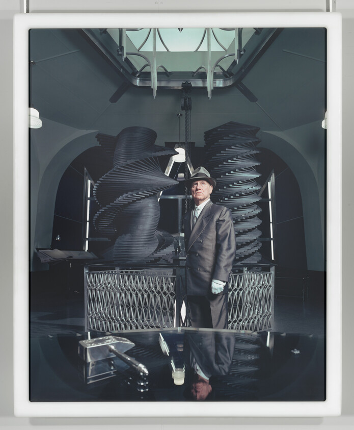 An older man in a suit and hat stands beside large spiral industrial machinery in a gallery.