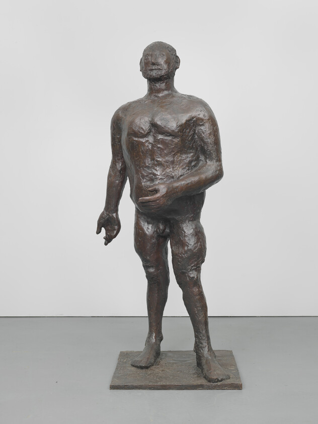 A rough-textured bronze sculpture of a standing nude male with one hand slightly extended.