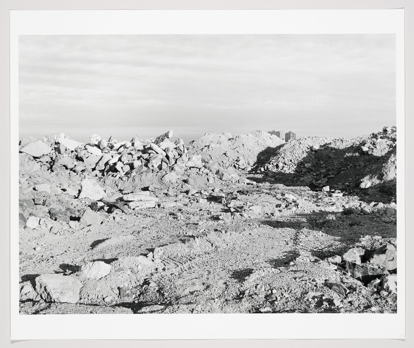 A barren construction site with large rubble piles and scattered rocks under a cloudy sky.