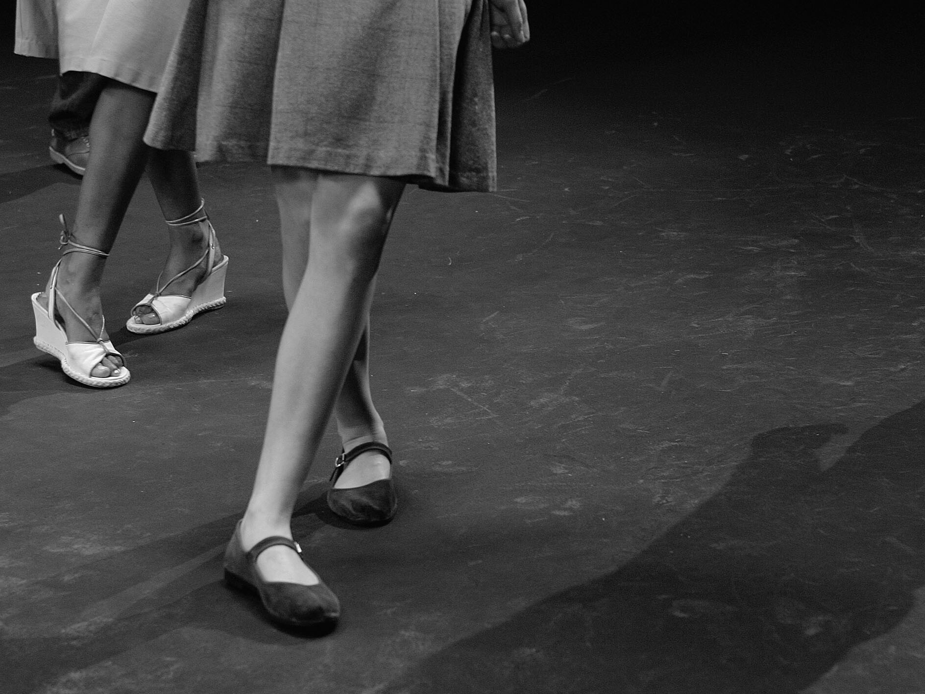 Two women walk on a dark runway wearing skirts, one in flats and one in wedge sandals.