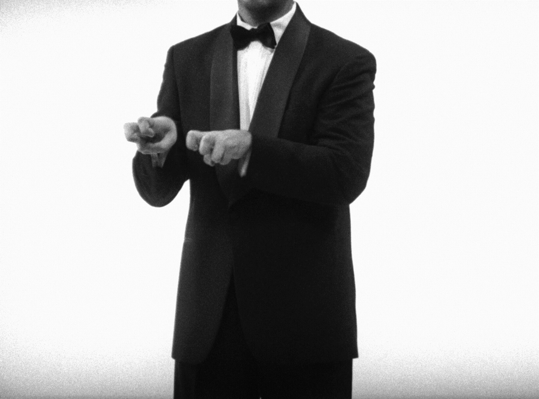 A man in a tuxedo points both hands forward with index fingers extended.