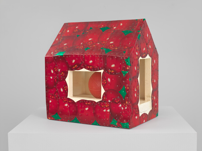 A small house-shaped sculpture covered in apple print with two cutout windows and a basketball inside.
